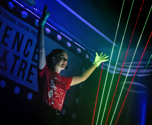 Live science show photo showing a female presenter with her hands in the air showing off the beams of light from a laser harp