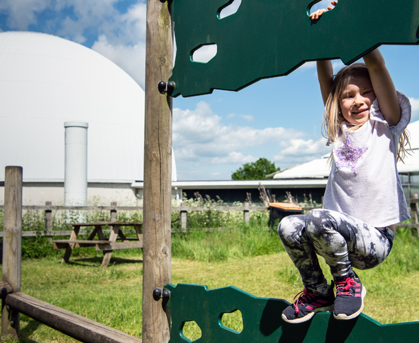 A girl plays on outdoor play equipment with the Planetarium dome behind her