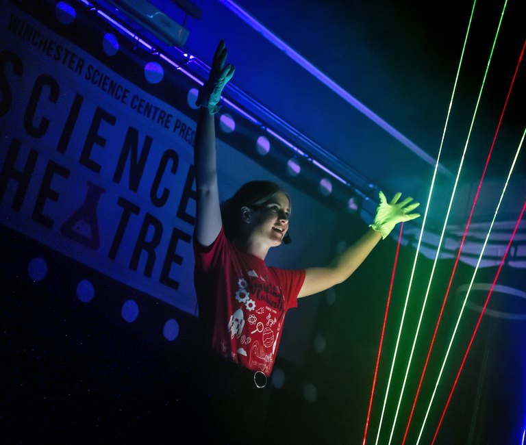 Live science show photo showing a female presenter with her hands in the air showing off the beams of light from a laser harp