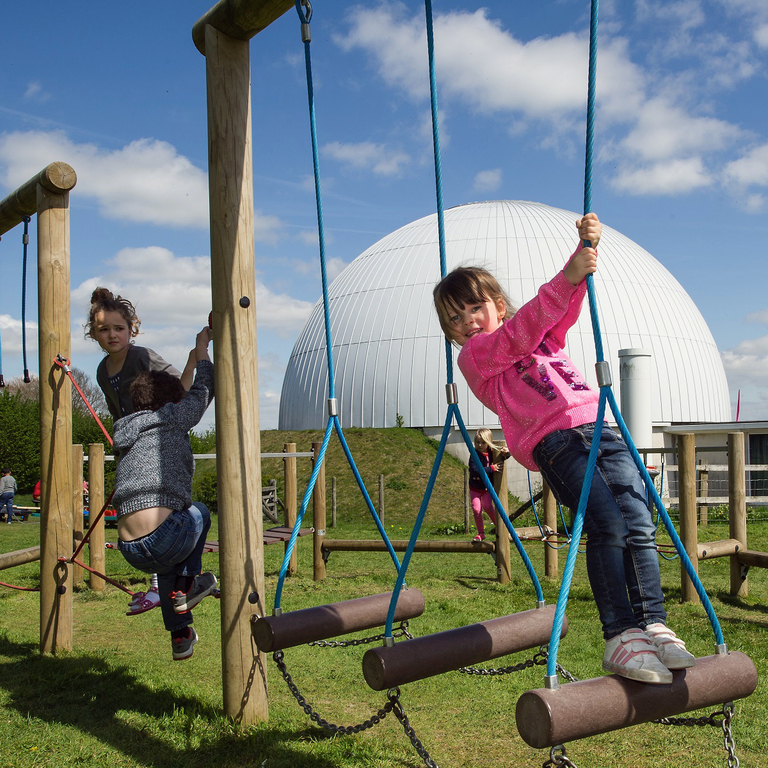 Children play on outdoor play equipment with the Planetarium dome behind them