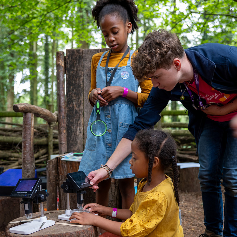 Two children and a science communicator in a forest looking at microscopes