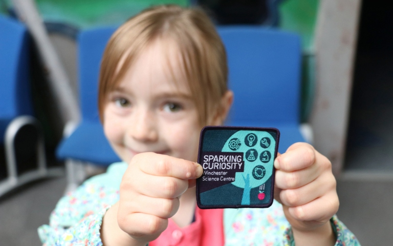 A young girl holding up her Curiosity Badge to the camera and smiling