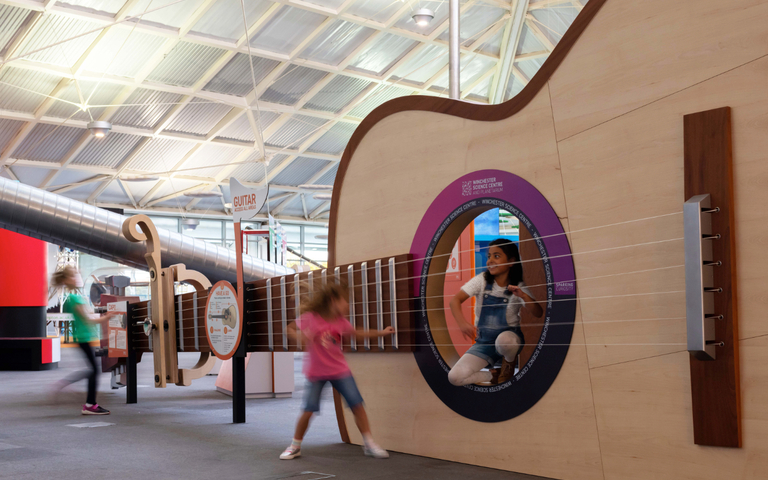 Interactive science exhibition photo showing two girls playing with a giant guitar