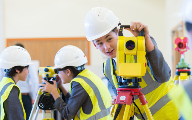 A school student wearing a hard hat and high visibility jacket uses a piece of equipment
