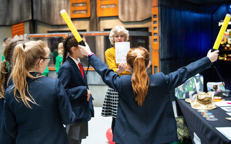 Girls in school uniform facing away from the camera holding yellow rods in the air, looking at what a teacher is holding up
