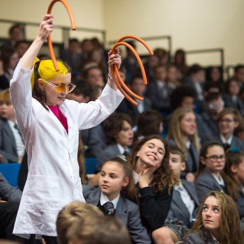 A presenter in a lab coat and safety goggles standing amongst a class of school children holding some rubber tubes up in the air