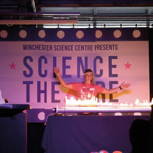 Live science show photo showing a female presenter dancing behind a tube lit up with flames