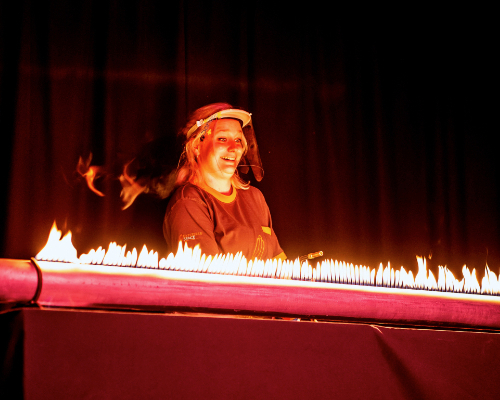 Live science show photo showing a female presenter smiling behind a tube lit up with flames