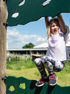 A smiling young girl hanging from part of the adventure playground outside