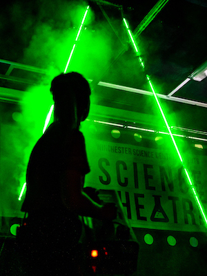 Live science show photo showing a presenter looking behind her at two green laser beams surrounded by smoke