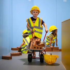 Interactive science exhibition photo showing a child in a high vis jacket and hard hat, pushing a small wheelbarrow of foam bricks