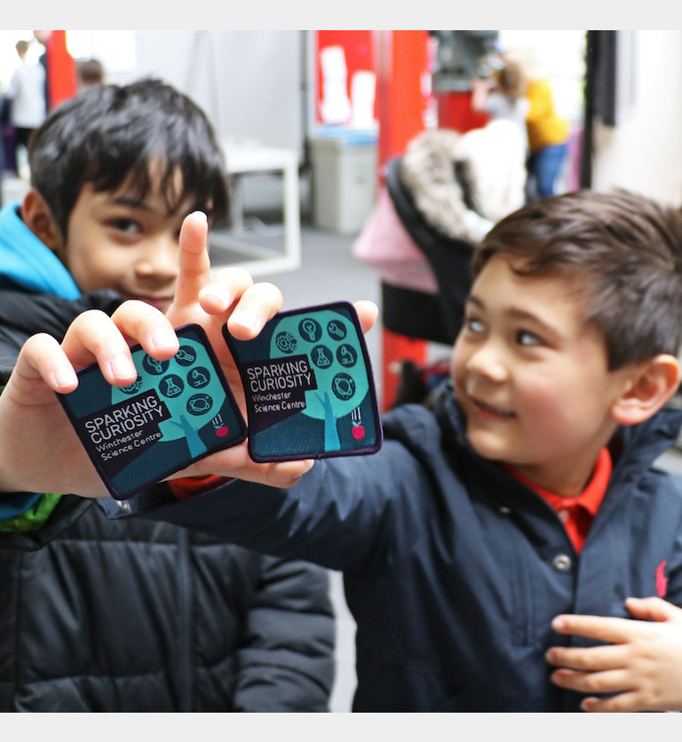 Two young boys holding up their Curiosity Badges to the camera and smiling