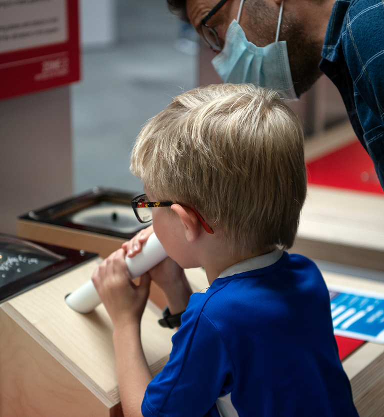 Interactive science exhibition photo showing a young boy with glasses blowing into a tube as part of an exhibit, with his parent standing behind