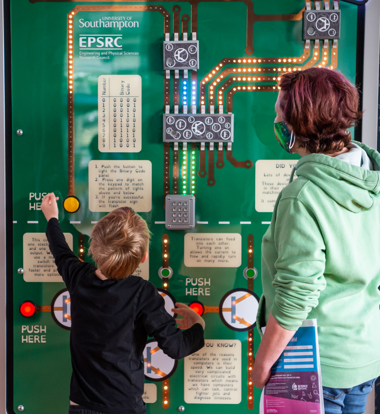 A boy and his mother play with a wall covered with buttons and cables