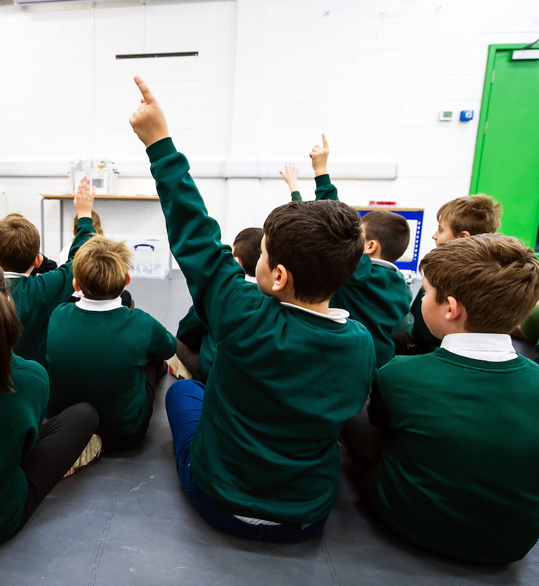 The backs of a group of school children sitting on the floor in a classroom, some with their hands in the air