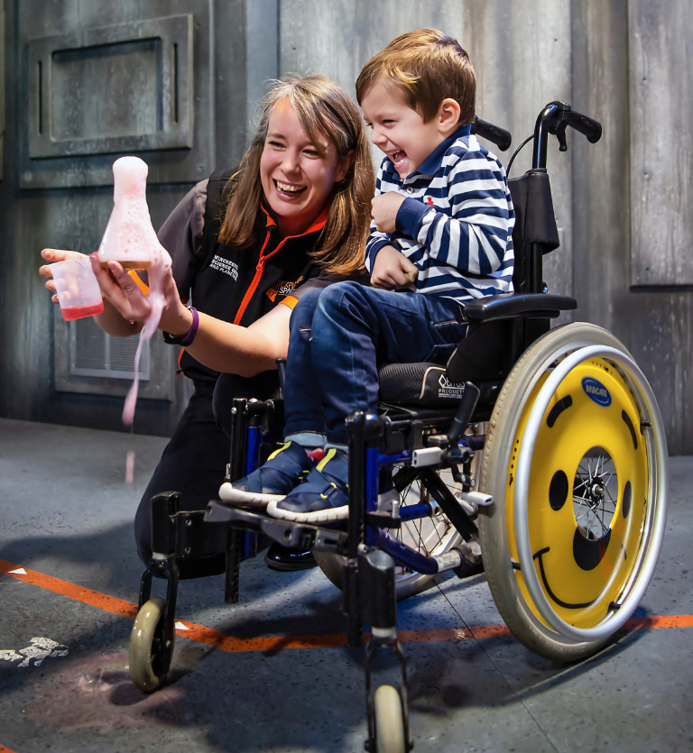 A young boy laughing in a wheelchair with a smiling presenter crouched beside him, holding a beaker containing a chemical reaction