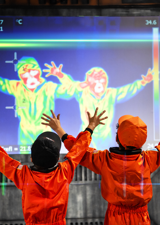 Interactive science exhibition photo showing two children in orange space suits and caps standing with their arms outstretched in front of a heat sensor screen