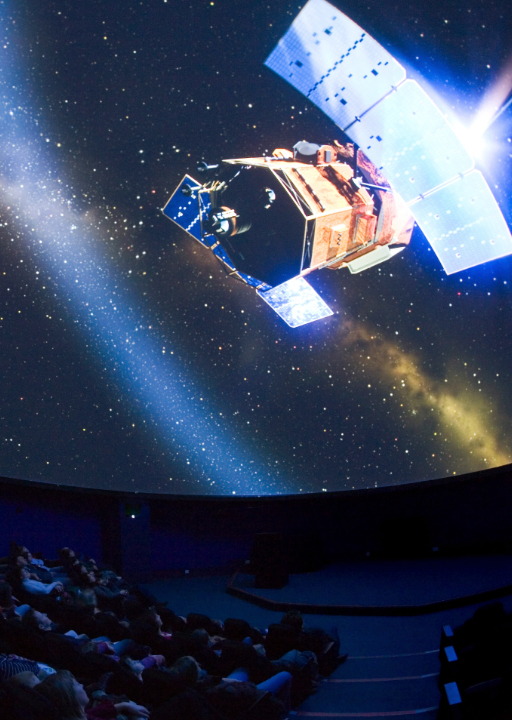 Planetarium photo showing an audience watching a satellite surrounded by stars being shown on the dome screen