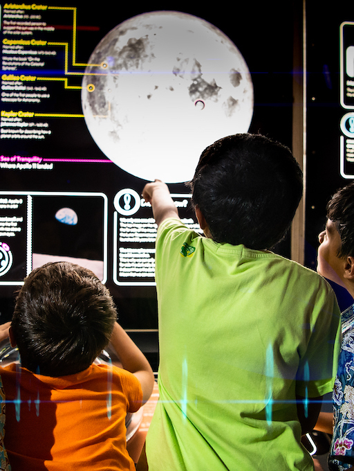 Interactive science exhibition photo showing two children in orange space suits and caps standing with their arms outstretched in front of a heat sensor screen